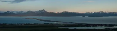 A panoramic shot of the Homer Spit stretching into Kachemak Bay, with the Kenai Mountains in the background at dusk.